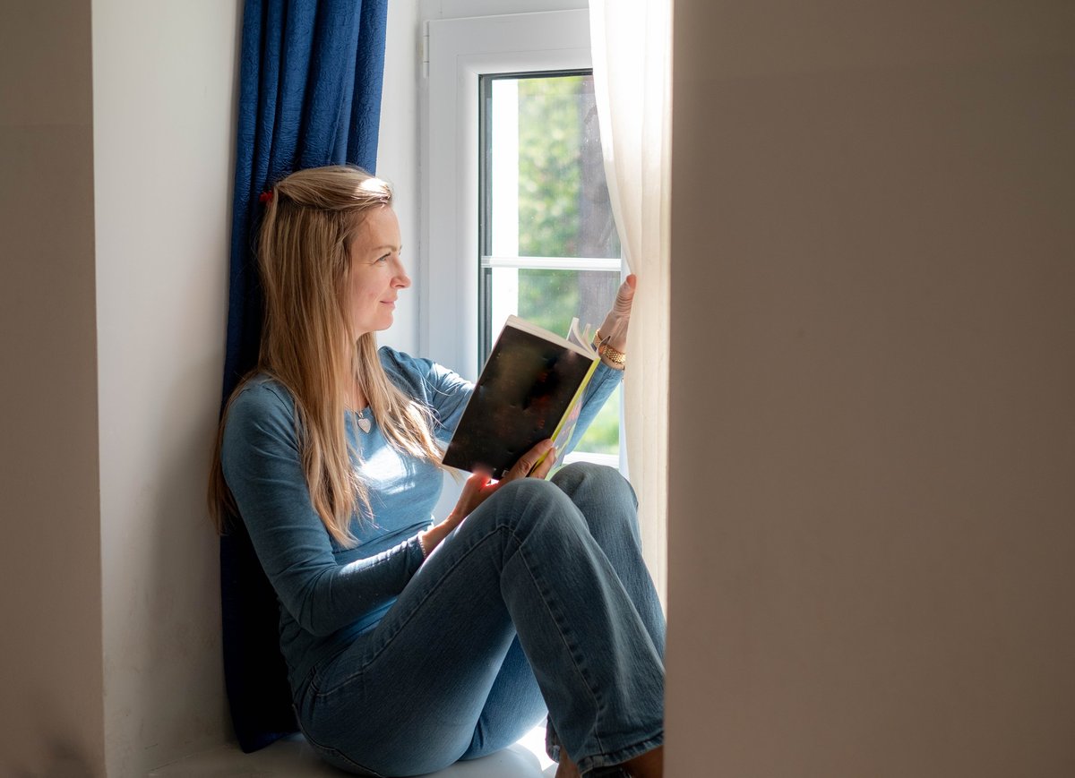 Woman enjoying a quiet moment reading a book while sitting by a bright window with soft natural light